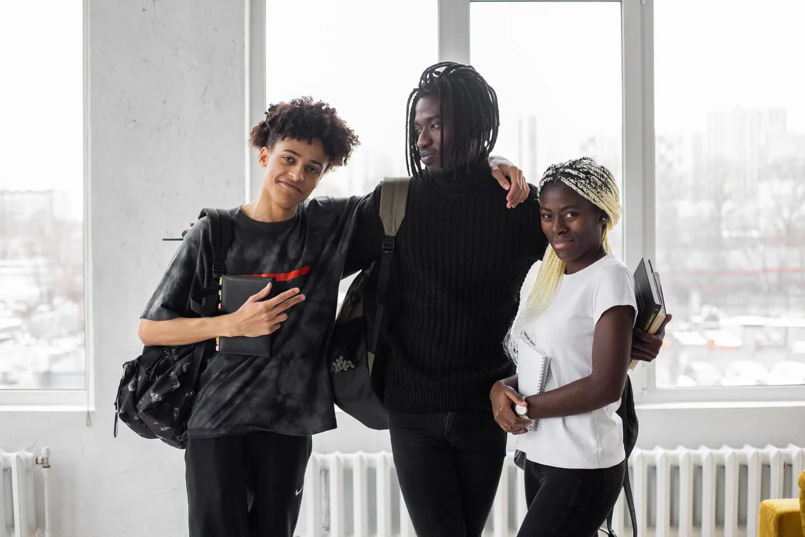 Trois étudiants souriants, sacs à dos et cahiers à la main, devant de grandes fenêtres lumineuses
